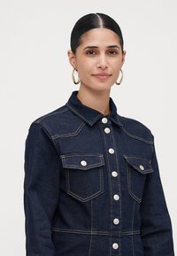 Γυναίκα με σκούρα μαλλιά πιασμένα πίσω, φορώντας χρ�υσά σκουλαρίκια κρίκους και μια σκούρα τζιν ζακέτα με κουμπιά και μπροστινές τσέπες, σε λευκό φόντο.