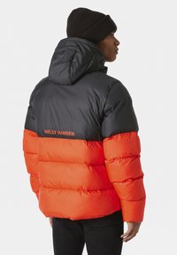 Giubbotto a vento con sezione superiore nera e parte inferiore arancione brillante, con trapuntatura orizzontale, cappuccio e logo del marchio sul retro.