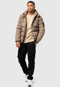 Hnedá puffová bunda so zipsom a kapucňou, skombinovaná s béžovým sweatshirtom a čiernymi cargo nohavicami. Biely sneakers dopĺňajú vzhľad.