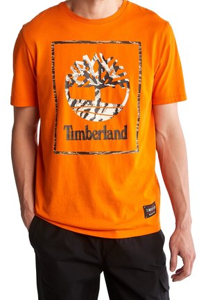 Mies, joka pukeutunut kirkkaan oranssiin Timberland-paitaan, jossa on leopardinraidallinen puulogo, ja mustiin housuihin, kädet taskuissa.