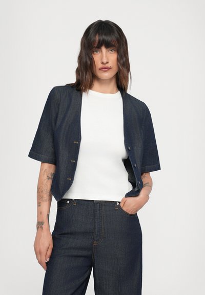 Giacca di jeans scura con maniche corte, chiusura a bottoni e un fit rilassato, indossata sopra un top bianco aderente. Pantaloni di jeans sartoriali completano l'outfit.