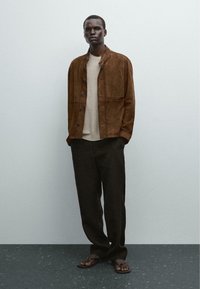 Veste en daim marron avec fermeture à boutons, sweater beige, pantalon noir et tongs marron. Textures lisses et palette de couleurs neutres.