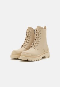 Botas de tobillo beige con cordones, hechas de material sintético suave y con una suela gruesa, que cuentan con una cremallera lateral y ojales de metal.