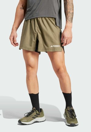 Pantalones cortos de color verde oliva con una capa inferior negra, cinturilla elástica y el logo "TERREX". Llevados con calcetas negras y zapatillas de correr oliva con detalles de malla.