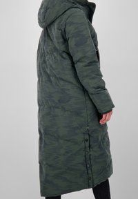 Lång grön puffjacka med huva, med en strukturerad mönster, dragkedjefickor på sidorna och flera knappar vid fållen.