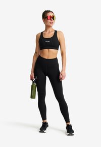Fit kvinna i svart sport-bh och leggings håller en grön vattenflaska, bär svarta löparskor och reflexglasögon, står på vit bakgrund.