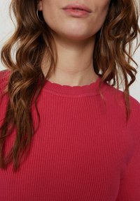 Ryša megzta apykaklė su iškirpte ir skersai susiūta šviesiai rožinė. Minkšta tekstūra ir prigludusi forma. Banguoti plaukai iš dalies matomi.