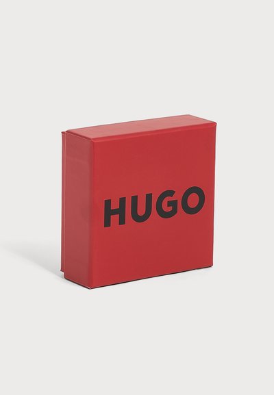 Червена правоъгълна кутия с удебелен черен текст "HUGO", центриран отпред, на обикновен светлосив фон.