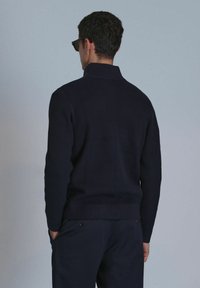 Marineblaue gerippte Sweater mit hohem Kragen, strukturiertem Stoff und tailliertem Schnitt. Trägt schwarze Hosen und Sonnenbrillen, zeigt die Rückansicht.