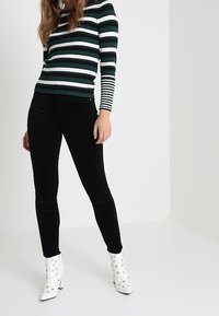 Vrouw in zwarte skinny broek, gestreepte lange mouwtop en witte enkellaarzen met gaatjesdetails, staande tegen een effen lichte achtergrond.