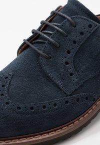 Chaussure habillée en daim bleu marine avec détails brogue, bout arrondi et cinq œillets. Dotée d'une semelle marron imitation cuir et de surpiqûres.