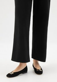 Ballerine in suede nero con dettaglio a barra in metallo dorato a forma quadrata, indossate con pantaloni larghi neri che mettono in mostra un tessuto texturizzato.
