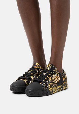 Schwarze Sneaker mit glänzendem Finish, gelbem Blumenmuster und schwarzer Gummikappe an der Spitze. Verfügen über schwarze Schnürsenkel und ein Logosymbol an der Seite.