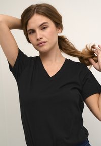Černé tričko s krátkými rukávy a výstřihem do V z hladké bavlněné tkaniny. Má volný střih a jednoduchý design. Modelka má vlasy v culíku.
