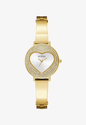 Aranyszínű női karóra szív alakú számlappal, kristályokkal díszített lünettával és arany színű mutatókkal, fehér számlapon, "GUESS" márkajelzéssel.