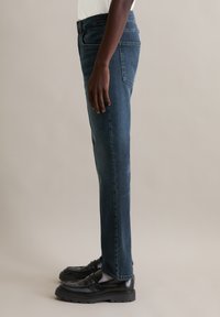 Jean en denim bleu foncé à coupe droite, présentant un design classique à cinq poches et des détails de couture minimalistes. Associé à des chaussures noires.