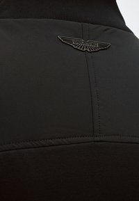 Tissu noir avec coutures cousues et un petit logo noir d'Aston Martin avec des ailes en haut au centre.