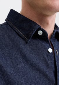Mørkeblå denimskjorte med knapp-up krage, hvite knapper og kontraststing. Nærbilde viser tekstur i stoffet og detaljene på kragen.