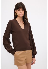 Brauner Strickpullover mit V-Ausschnitt und strukturierten Ballonärmeln. Kombiniert mit hellbraunen Leder-Shorts, die einen modernen Lagenlook unterstreichen.