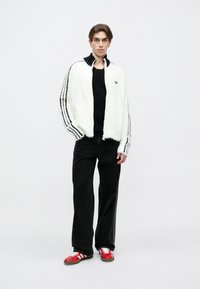 Chaqueta blanca difusa con cuello negro y tres rayas negras, combinada con pantalones anchos negros y zapatillas rojas con detalles blancos.