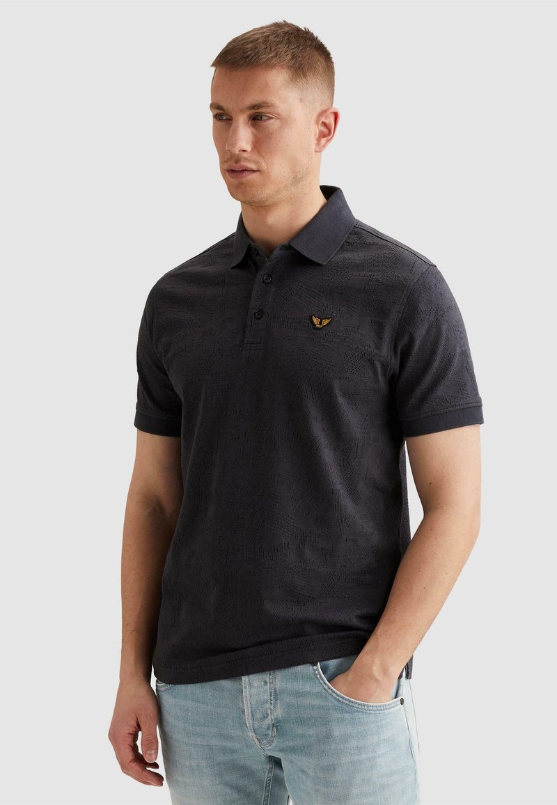 Homme portant un polo à manches courtes à motif sombre et un jean bleu clair, avec une main dans la poche, regardant légèrement vers la gauche.
