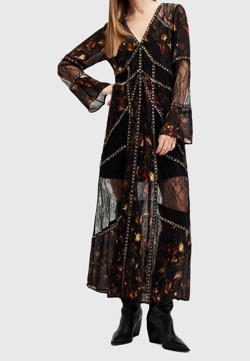 Femme portant une longue robe noire avec des motifs floraux orange, des panneaux en dentelle transparente, une bordure décorative, des manches évasées et des bottes noires pointues.