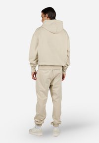 Beige hettegenser og matchende joggebukser, laget av myk stoff. Har hette, ribbede mansjetter og smalnede ben. Brukt med hvite sneakers.