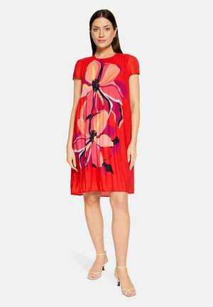 Woman wearing knee-length red dress with large pink and orange floral print, short sleeves, and beige heeled sandals, standing on white background.