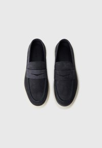 Pár tmavě námořnických semišových loafers s bílými podrážkami a dekorativním šitím, uspořádaných vedle sebe na bílém pozadí.
