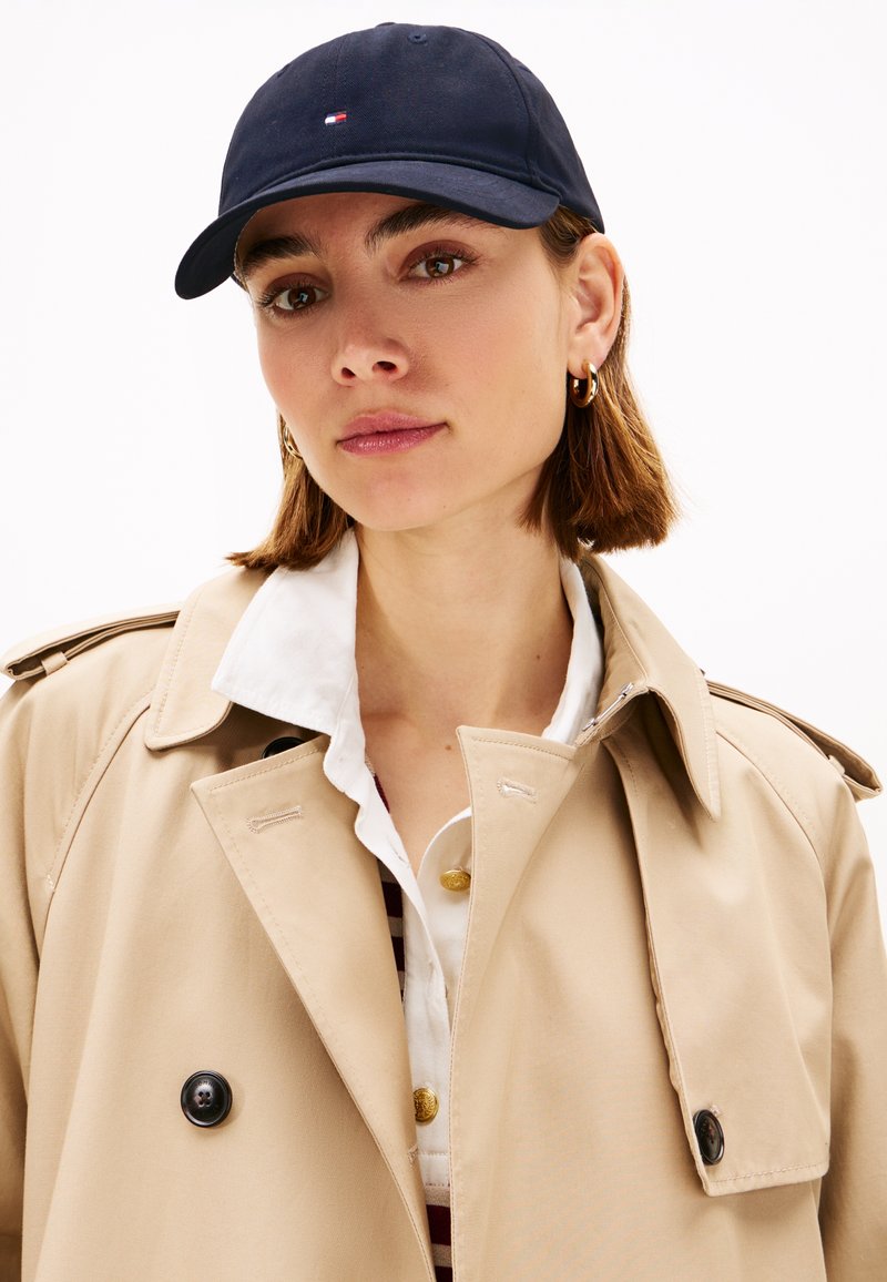 Casquette de baseball bleu marine en coton avec un logo à l'avant. Portée avec un trench-coat beige et une chemise blanche à col, présentant un détail de bouton.