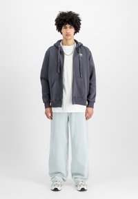 Γκρι φούτερ με μπροστινές τσέπες, φερμουάρ και κορδόνια, φορεμένο πάνω από ένα λευκό μπλουζάκι. Συνδυασμένο με ανοιχτό μπλε φαρδύ τζιν και αθλητικά παπούτσια.