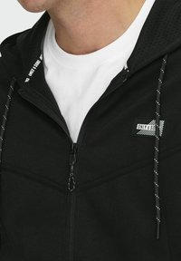 Hoodie zippé noir avec une capuche perforée, des cordons blancs, et un patch logo. T-shirt blanc visible sous le col.