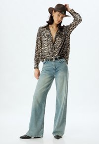Chemise en soie à imprimé léopard, jeans en denim bleu clair larges, bottes noires à bout pointu et un chapeau en feutre marron avec un band décoratif.