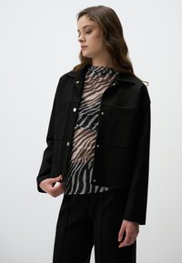 Zwarte cropped jas met gouden drukknoopjes, oversized zakken, gedragen over een doorzichtige top met een zebra-print en subtiele glittertjes.