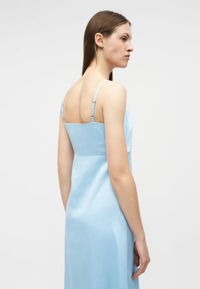 Γυναίκα με ίσια καστανά μαλλιά που φοράει ένα ανοιχτό γαλάζιο σατέν φόρεμα με λεπτές ρυθμιζόμενες τιράντες, εμφανιζόμενη από την πίσω και πλάγια πλευρά.
