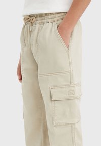 Beżowe spodnie cargo z elastycznym paskiem w talii i sznurkiem, bocznymi kieszeniami oraz kieszenią na udzie, noszone przez osobę z ręką w kieszeni.