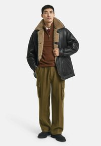Schwarze Lederjacke mit Lammfellkragen, brauner Pullover und olivfarbene Cargo-Hose. Schwarze Schuhe vervollständigen das Outfit.