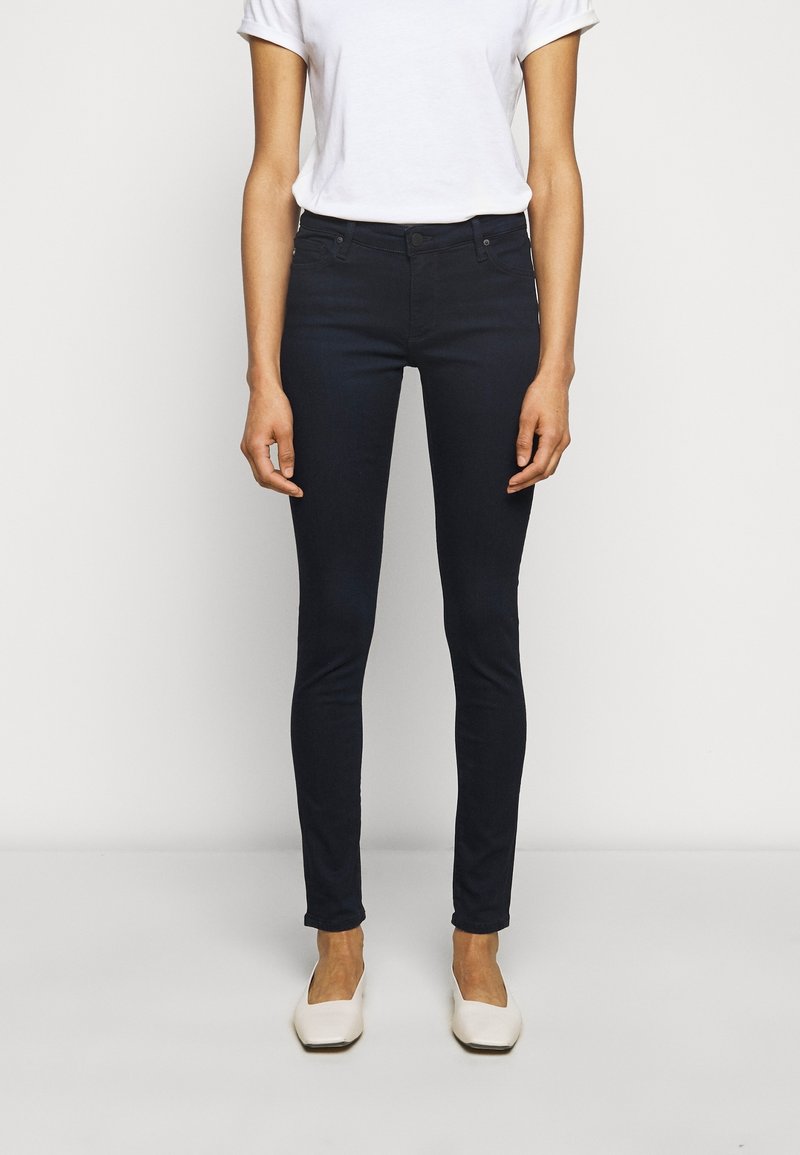 Personne portant un jean skinny noir, une chemise blanche à manches courtes et des chaussures blanches à bout carré, debout devant un fond clair uni.