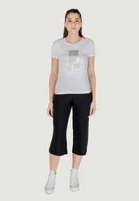 Camiseta blanca de manga corta con un estampado gráfico metálico, combinada con pantalones negros cortos y zapatillas altas blancas. Diseño simple y casual.