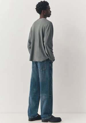 Mladý muž stojící bokem, oblečený v šedém tričku s dlouhým rukávem, volných modrých džínách a černých botách, na pozadí jednoduché bílé stěny.