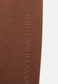 Tecido canelado castanho com texto em relevo "BRUUNS BAZAAR". A textura é suave com linhas verticais, apresentando um design simples e moderno.