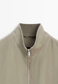 Veste beige avec col haut et fermeture éclair argentée sur le devant, en tissu texturé.