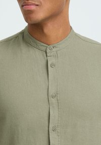 Chemise vert olive clair en tissu texturé, avec un col mandarin et une fermeture à boutons assortis et petits.