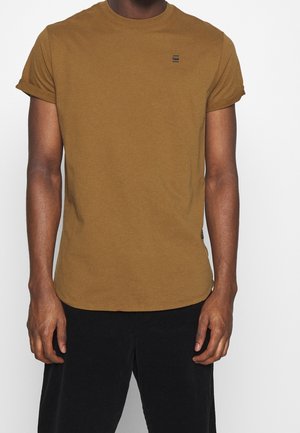 Man draagt een bruin T-shirt met korte mouwen die zijn opgerold en zwarte broek, staand tegen een effen witte achtergrond.
