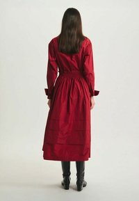 Robe rouge mi-longue avec une taille cintrée, des manches longues, un ourlet plissé et un tissu texturé. Associée à des bottes noires jusqu'au genou.