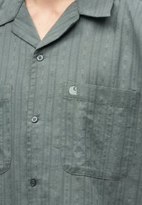 Γκρι πουκάμισο με κουμπιά και υφή κάθετων ριγών, με δύο τσέπες στο στήθος, η μία με μικρό κεντητό λογότυπο.