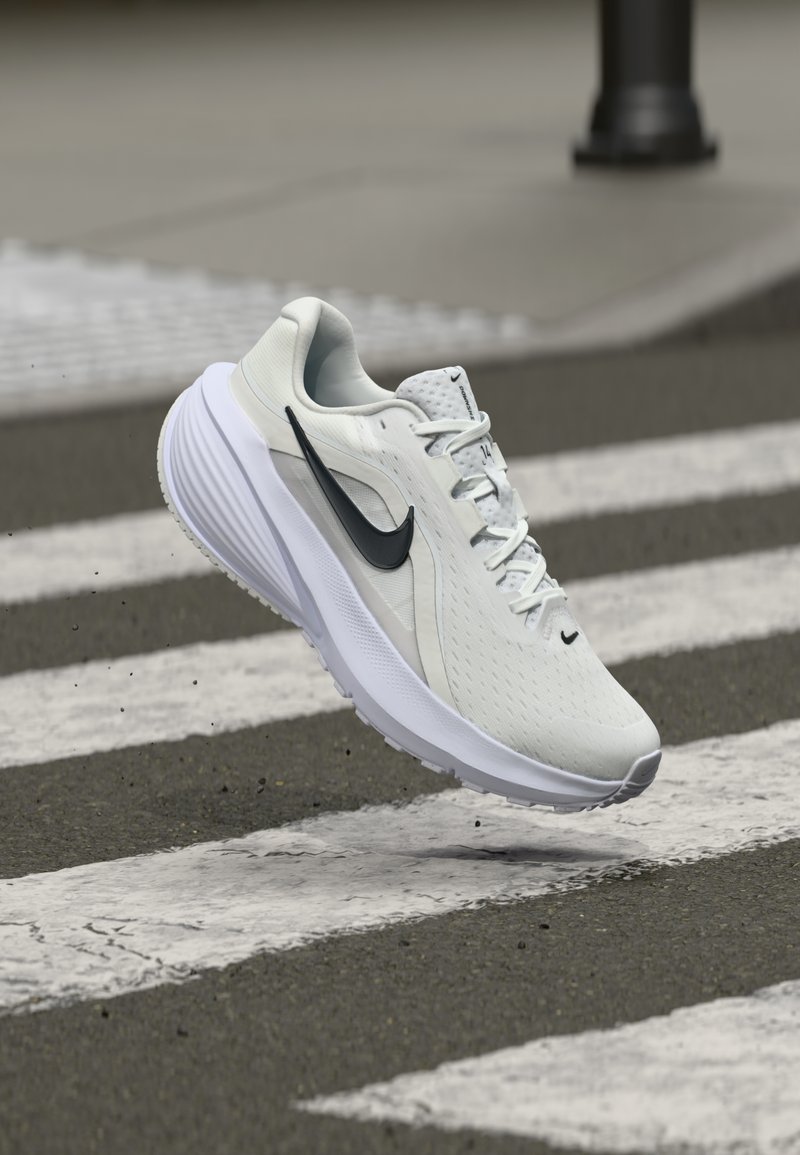 Zapatilla de correr Nike en gris claro con swoosh negro, parte superior texturizada, collar acolchado, acentos reflectantes y suela blanca, mostrada en el aire sobre un paso de peatones.