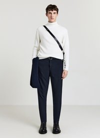 Weißer, gerippter Rollkragenpullover, marineblaue, gemusterte Hose, schwarzer Gürtel, schwarze Stiefeletten und ein strukturierter, marineblauer Schal, der über dem Arm drapiert ist.