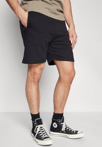Shorts en coton noirs avec une coupe décontractée, dotés de poches latérales et d'une texture lisse, associés à des baskets montantes noires et des chaussettes noires.