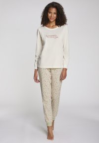 Crème lange mouwen top met de tekst "Cozy", gecombineerd met groene broek met een rood zuurstokpatroon. Zachte stof, ontspannen pasvorm, elastische manchetten.